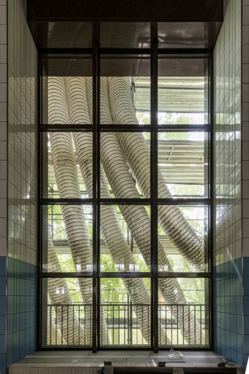 Grosse Fensterfront eingerahmt von einer gefliesten Wand mit Blick auf mehrere flexible Lüftungsrohre, die sich von einem Gerüst nach unten wölben. Draussen sind ein Baugerüst und grüne Bäume zu sehen. 